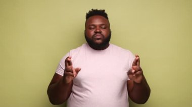 Umut dolu Afro-Amerikalı adam pembe tişört giyiyor ve gözlerini kapatıyor. Dilek diliyor, mucizeye inanıyor, başarıya inanıyor. Açık yeşil arka planda kapalı stüdyo çekimleri.