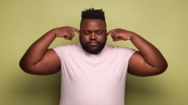 Bunu daha fazla duymak istemiyorum. Pembe tişört giyen, parmaklarını kulağına dayayan, rahatsız edici sesler çıkaran Afro-Amerikan bir adam. Açık yeşil arka planda kapalı stüdyo çekimleri.