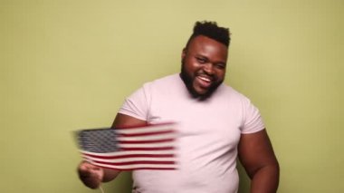 Pembe t-shirt giyen Afro-Amerikalı bir adam elinde ABD bayrağı ile kameraya bakarken dişlek gülümseme, insan hakları, konuşma özgürlüğü. Açık yeşil arka planda kapalı stüdyo çekimleri.