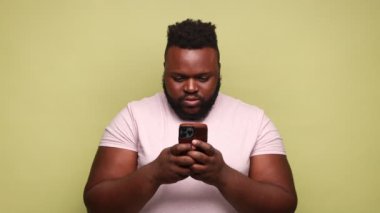 Şaşırmış Afro-Amerikalı bir adam pembe tişört giyiyor cep telefonu kullanıyor, sosyal ağları kontrol ediyor, internete bakıyor. Açık yeşil arka planda kapalı stüdyo çekimleri.