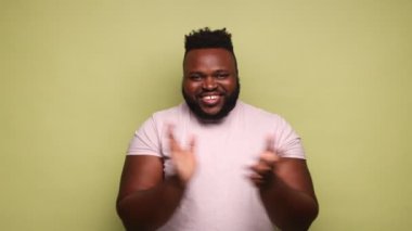 Neşeli sakallı Afro-Amerikalı adam pembe tişört giyiyor, el çırpıyor, performansını alkışlıyor, mutlu görünüyor, bravo. Açık yeşil arka planda kapalı stüdyo çekimleri.
