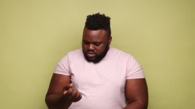 Mutsuz, hasta, sakallı, pembe tişört giyen, acı çeken, travma geçiren, bileğini burkan, ağrıyan Afro-Amerikan bir adam. Açık yeşil arka planda kapalı stüdyo çekimleri.