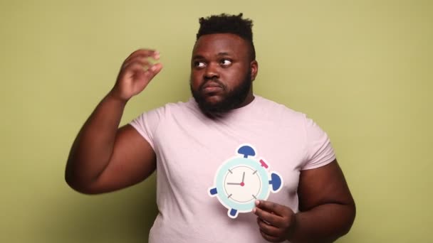 ピンクのTシャツを着たアフリカ系アメリカ人の男性は 紙の目覚まし時計を持っていて 締め切りについて考え 頭をこすりました 薄い緑の背景に隔離された屋内スタジオショット — ストック動画