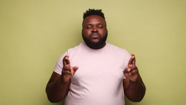ピンクのTシャツを着たアフリカ系アメリカ人男性が指を渡り 目を閉じて願いを叶え 奇跡を信じ 成功することを願っています 薄い緑の背景に隔離された屋内スタジオショット — ストック動画