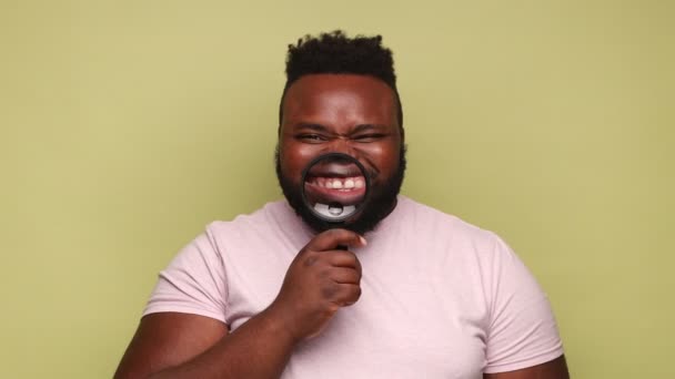 一个穿着粉色T恤 牙齿上挂着放大镜的有趣而积极的非洲裔美国男人 满面春风地看着相机 室内拍摄被隔离在浅绿色背景下 — 图库视频影像