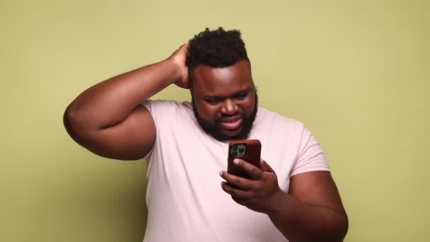 多愁善感的年轻非洲裔美国人 身穿粉色T恤 用手机思考着 揉揉着头 思考着答案 室内拍摄被隔离在浅绿色背景下 — 图库视频影像