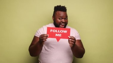 Yakışıklı, Afro-Amerikalı, pembe t-shirt tutan adam beni takip et ve bloguna abone olmak istediğini söyle. Açık yeşil arka planda kapalı stüdyo çekimleri.