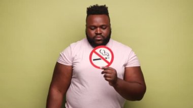 Ciddi bir Afro-Amerikan erkeği pembe tişört giyiyor ve elinde sigara içilmez tabelasına işaret ediyor, sigarayı bırakıyor, başını sallıyor. Açık yeşil arka planda kapalı stüdyo çekimleri.