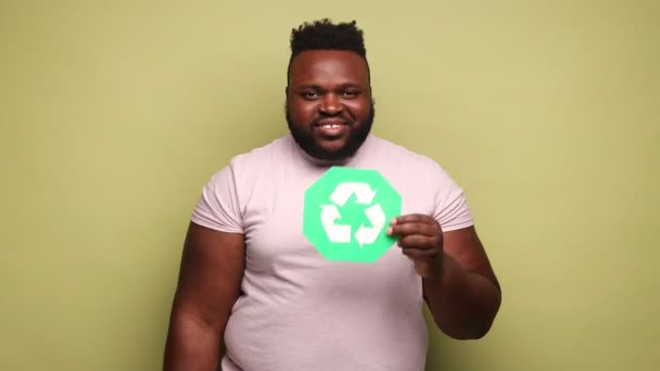 喜びに満ちた陽気なアフリカ系アメリカ人の男は ピンクのTシャツを着て緑のリサイクルサインを保持し 親指を表示し カメラに指を指す 薄い緑の背景に隔離された屋内スタジオショット — ストック動画