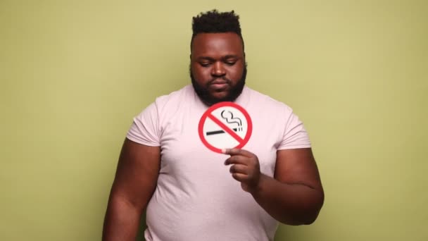深刻なアフリカ系アメリカ人の男性はピンクのTシャツを保持し 手の中に喫煙記号を指して いいえうなずいて禁煙を着て 薄い緑の背景に隔離された屋内スタジオショット — ストック動画