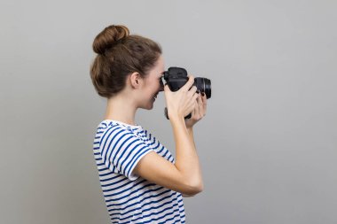 Kadın fotoğrafçı ya da gezginin çizgili tişört giyip elinde profesyonel dijital dslr kamera tutarken, odaklanırken, fotoğraf çekerken ya da video çekerken yan görüntüsü. Gri arka planda izole edilmiş kapalı stüdyo görüntüleri.