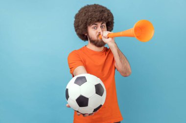 Afro saçlı, turuncu tişörtlü favori futbol takımının zaferini kutlayan, elinde boynuz ve top olan bir adamın portresi. Mavi arka planda kapalı stüdyo çekimi.