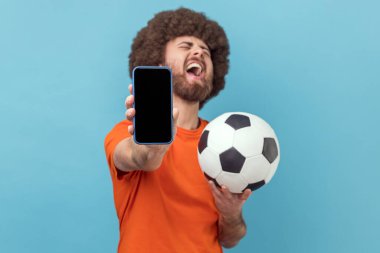 Son derece mutlu, Afro saçlı, tişörtlü futbol topuyla ayakta duran, cep telefonunu boş gösteren, bahis oynayan ve kazanan bir adam. Mavi arka planda kapalı stüdyo çekimi.