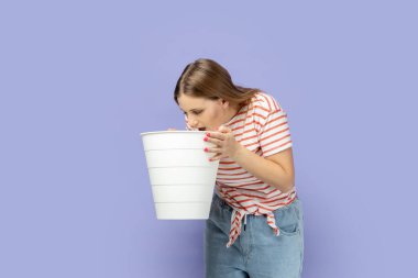 Çizgili tişört giyen sarışın bir kadının portresi ani mide ağrısı, mide bulantısı ve kusmuk çekiyor, elinde çöp kovası, hazımsızlık. Mor arka planda izole edilmiş kapalı stüdyo çekimi.