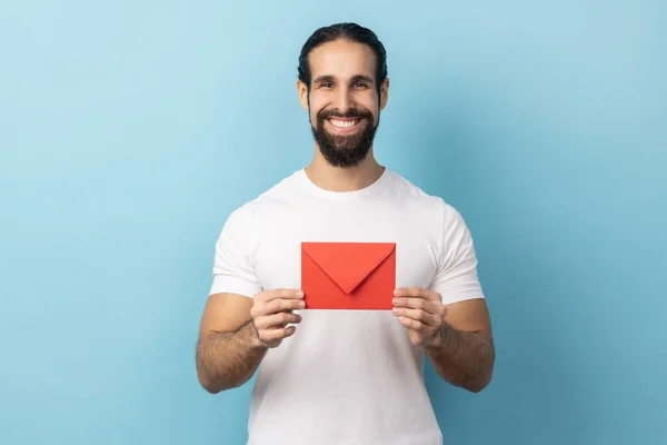 I got love letter on Valentine\'s day. Portrait of man with beard in white T-shirt holding letter in red envelope or greeting card and smiling joyfully. Indoor studio shot isolated on blue background.