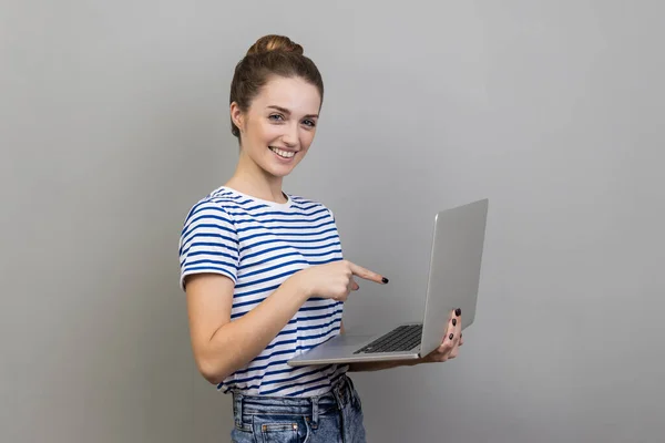 ストライプのTシャツを着て ラップトップを保持し 画面を指している女性の肖像画は コンピュータアプリケーション インターネットニュースについて満足した 灰色の背景に隔離された屋内スタジオショット — ストック写真