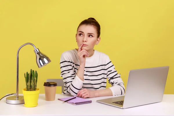 穿着条纹衬衫的多愁善感的女人坐着 一边在办公室的笔记本电脑上工作 一边沉思着新项目 在黄色背景下隔离的室内工作室拍摄 — 图库照片