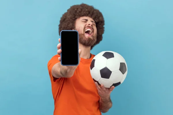 非常快乐的黑人发型男人穿着T恤 站在足球旁边 拿着空的展示手机 投注和赢球 室内拍摄被蓝色背景隔离 — 图库照片