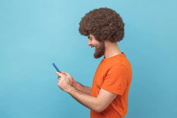 オレンジ色のTシャツを着て スマートフォンを使用して 幸せな笑顔の顔を持つビデオを見て アフロの髪型と肯定的な男性ブロガーのサイドビュー 青い背景に隔離された屋内スタジオショット — ストック写真