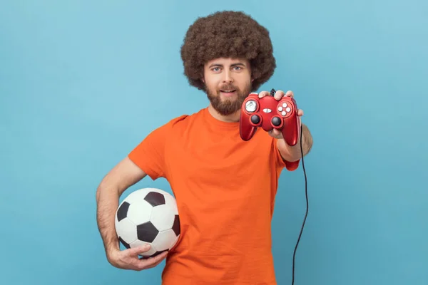 英俊的黑人男子穿着橙色T恤 伸出操纵杆和足球球 提供虚拟游戏的形象 室内拍摄被蓝色背景隔离 — 图库照片
