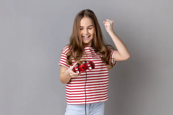 ストライプのTシャツを身に着けている少女の肖像画手赤ジョイスティックパッドを保持し 心から喜び オンライン仮想ゲームで勝利イエスジェスチャーを示しています 灰色の背景に隔離された屋内スタジオショット — ストック写真