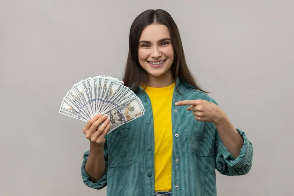 喜んで笑顔の女性はドル紙幣のファンを指して 幸せな表情でカメラを見て カジュアルなスタイルのジャケットを着ている 灰色の背景に隔離された屋内スタジオショット — ストック写真
