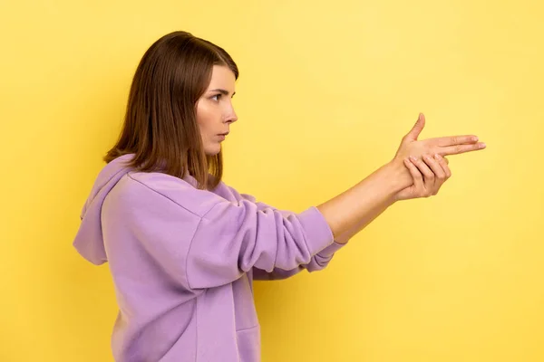 あなたを殺すわ 怒っている女性の側のビューのカメラに指銃を指して 撮影すると 手の武器を模倣し 紫色のパーカーを着て脅迫 黄色の背景に隔離された屋内スタジオショット — ストック写真