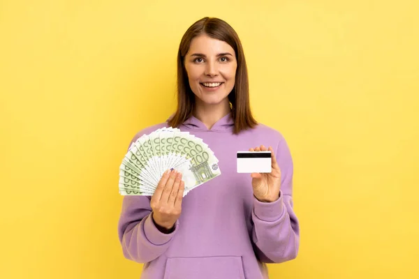 ユーロ紙幣やクレジットカードのファンを保持暗い髪を持つ肯定的な女性の笑顔の肖像画 お金を稼ぐ 紫色のパーカーを着て 黄色の背景に隔離された屋内スタジオショット — ストック写真