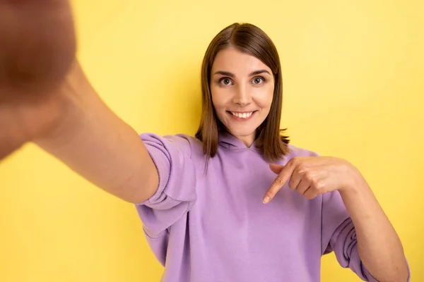 Smiling attractive woman taking selfie, point of view of photo, pointing finger down, recommend to subscribe her vlog, wearing purple hoodie. Indoor studio shot isolated on yellow background.