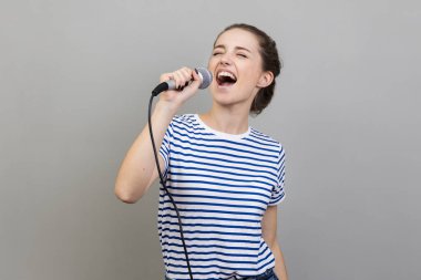 Portrait of excited positive woman wearing striped T-shirt singing songs at karaoke, having fun, holding microphone, entertainment. Indoor studio shot isolated on gray background. clipart