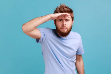 Portrait of serious concentrated bearded man looking far away at distance with hand over head, attentively searching for bright future. Indoor studio shot isolated on blue background. clipart