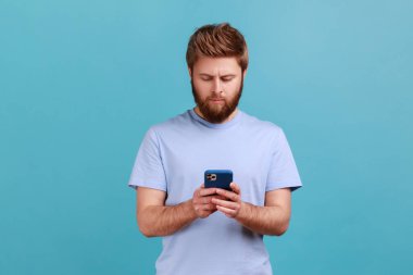 Ciddi derecede konsantre sakallı bir adamın portresi cep telefonuyla sosyal medyada mesaj yolluyor, cep telefonu servislerini kullanıyor, internette sohbet ediyor. Mavi arka planda kapalı stüdyo çekimi.