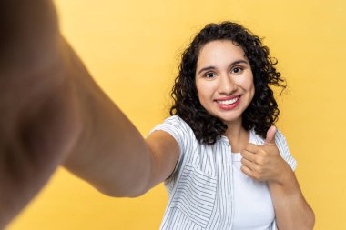 Çekici, iyimser, çekici bir kadının portresi koyu renk dalgalı saçlı, bakış açısı olan, jest, baş parmak yukarı, iyi geri bildirim, POV gibi fotoğraflar yapan. Sarı arka planda kapalı stüdyo çekimleri.