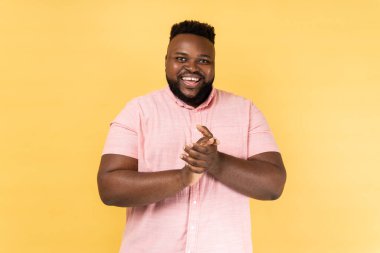 Pembe tişört giyen, avuçlarında alkışlayan, alkışlayan ve zaferle tebrik eden olumlu arkadaş canlısı bir adamın portresi. Sarı arka planda kapalı stüdyo çekimleri.