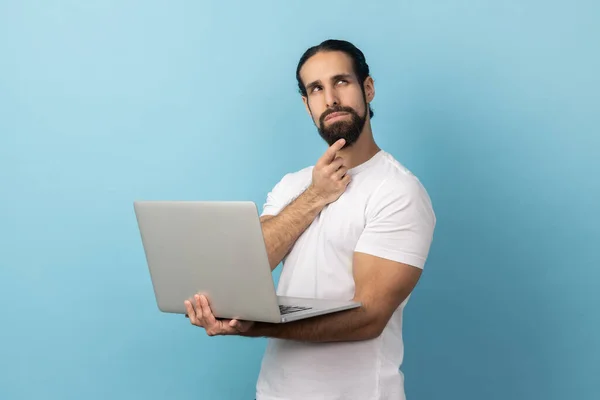 白いTシャツを着た髭を生やした男の肖像画がノートパソコンを持っていて 新しいビジネスプロジェクトを考えて離れて見ている顎をこすります 青い背景に隔離された屋内スタジオショット — ストック写真