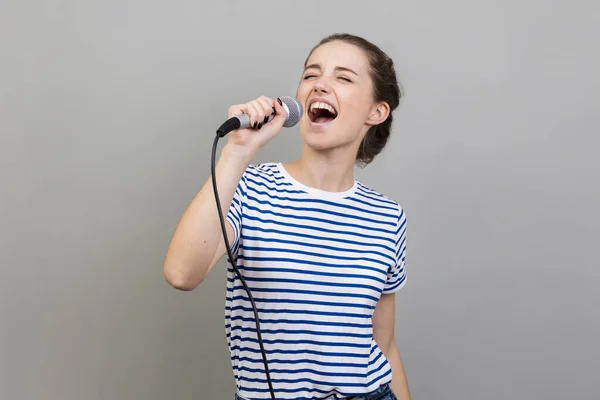 在卡拉Ok上穿着条纹T恤衫唱歌 玩的开心 拿着话筒 娱乐的兴奋而积极的女人的画像 在灰色背景下被隔离的室内拍摄 — 图库照片