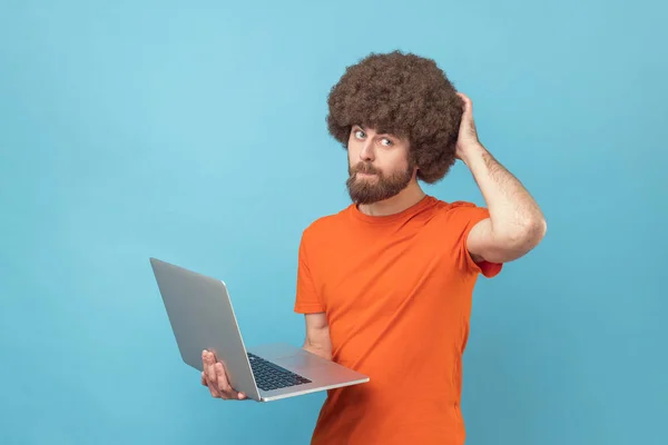 オレンジ色のTシャツを着たアフロヘアの男の肖像画は ノートパソコンを持ち 新しいプロジェクトについて考え 彼の頭の後ろを引っ掻いています 青い背景に隔離された屋内スタジオショット — ストック写真