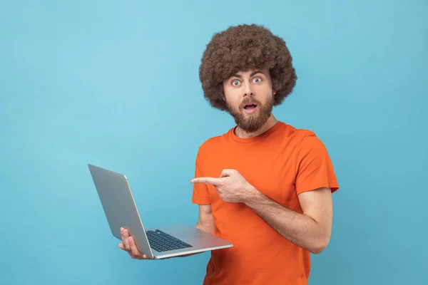穿着橙色T恤的非洲裔男子的画像 指着笔记本电脑屏幕 用震惊的表情 张开的嘴看着相机 室内拍摄被蓝色背景隔离 — 图库照片