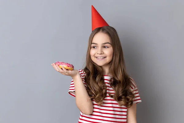 一个快乐快乐的小女孩穿着条纹T恤和派对锥 手握棕榈味甜甜甜圈 面带微笑地看着甜食的画像 在灰色背景下被隔离的室内拍摄 — 图库照片