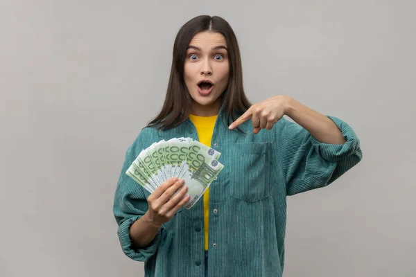 興奮した女性は 手にユーロ紙幣を指して 大きなお金を保持するために驚いている式を見て カジュアルなスタイルのジャケットを着て驚いている 灰色の背景に隔離された屋内スタジオショット — ストック写真