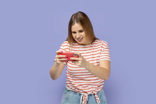 スマートフォンでゲームをプレイし 良い感情を表現し 彼女のゲームを楽しんで縞模様のTシャツを着て魅力的な肯定的なブロンドの女性のゲーマーの肖像画 紫色の背景に隔離された屋内スタジオショット — ストック写真