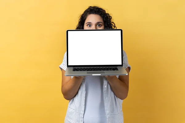害羞的女人的肖像 深色波浪般的头发 半张脸隐藏在笔记本电脑后面 带着空洞的显示 复制空间来促销 在黄色背景下隔离的室内拍摄 — 图库照片