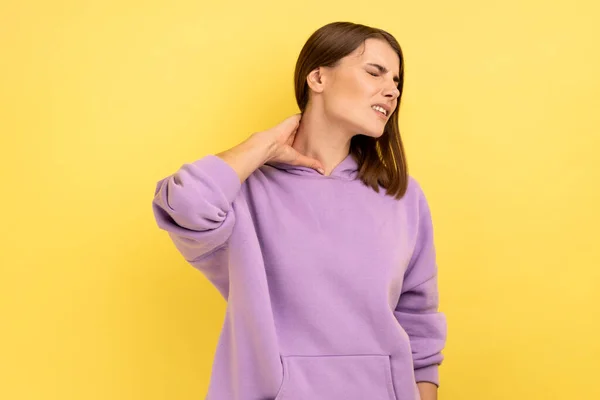 疲れ不健康な女性の肖像首の痛み 筋肉の緊張 子宮頸椎の先端神経 紫色のパーカーを着て苦しんでいます 黄色の背景に隔離された屋内スタジオショット — ストック写真