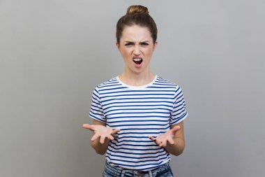 Çizgili tişört giyen, şok ve yanlış anlaşılmayla el ele tutuşan sinirli mutsuz bir kadının portresi. Nasıl imzalarsın? Gri arka planda izole edilmiş kapalı stüdyo görüntüleri.