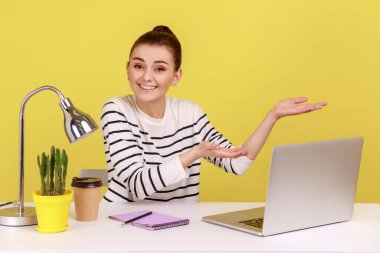 Ofiste çalışan mutlu bir kadının portresi. Masasında dizüstü bilgisayarla. Hoş geldin jesti yapıyor, müşteriyi selamlıyor. Sarı arka planda kapalı stüdyo çekimleri.