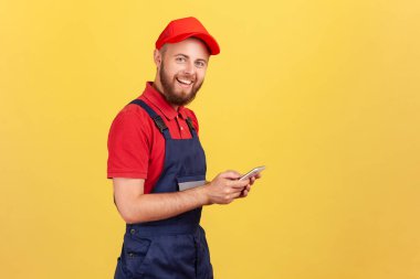 Cep telefonunda yazan mutlu işçi görüntüsü, cep telefonu kuryesini kullanarak çevrimiçi siparişi teslimat olarak kabul eden, gülümseyerek kameraya bakan. Sarı arka planda kapalı stüdyo çekimleri.