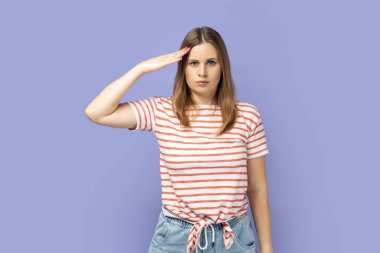 Responsible patriotic blond woman wearing striped T-shirt following discipline, saluting to commander with hand near temple and listening order. Indoor studio shot isolated on purple background. clipart