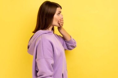 Şok olmuş, koyu renk saçlı bir kadının portresi. Elini ağzına koyup ileriye bakıyor, korkunç bir sır saklıyor, mor kapüşonlu bir şey giyiyor. Sarı arka planda kapalı stüdyo çekimleri.