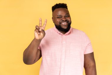 Pembe gömlekli bir adamın zafer jesti yaptığı ve kameraya şen şakrak baktığı, huzuru gösteren, çift parmaklı v işareti. Sarı arka planda kapalı stüdyo çekimleri.