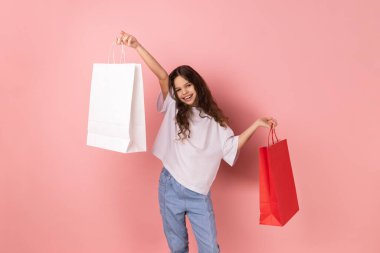 Beyaz tişört giyen, elinde kağıt çantalar olan mutlu küçük bir kızın portresi. Alışveriş merkezi indirimleri ve iyi alışverişlerle heyecanlanıyor. Pembe arka planda kapalı stüdyo çekimleri.